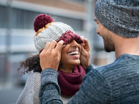 A person playfully pulls a knit cap over another person's eyes, both are smiling and wearing knit hats and sweaters in a casual outdoor setting.