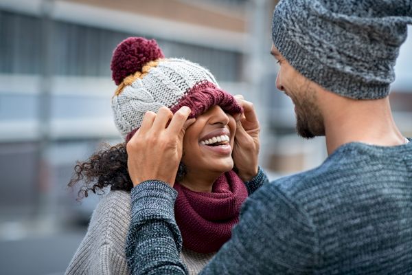 A person playfully pulls a knit cap over another person's eyes, both are smiling and wearing knit hats and sweaters in a casual outdoor setting.