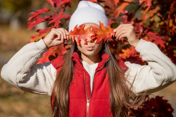 A person stands outdoors, holding vibrant orange autumn leaves over their eyes. They wear a white beanie, white sweater, and red vest.