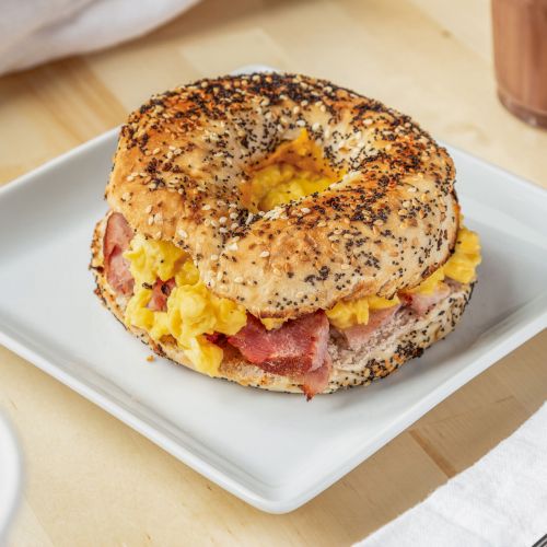 A bagel sandwich with scrambled eggs and bacon on a square white plate, with a fork and napkin nearby, and a drink and dessert in the background.