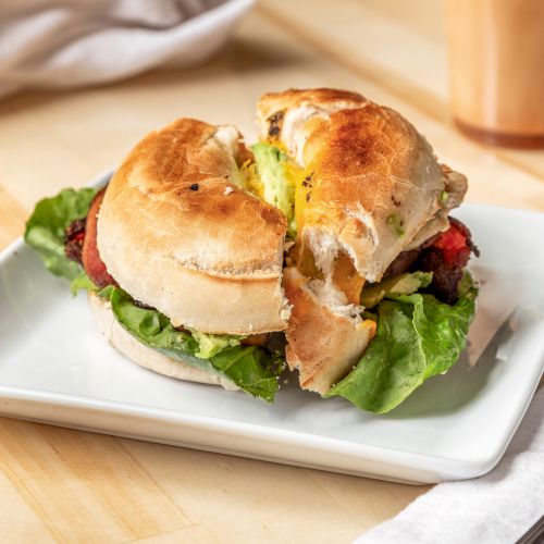 A bagel sandwich with lettuce and other fillings on a white plate, placed on a wooden table with a fork, napkin, and cup nearby.