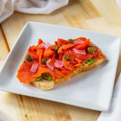 The image shows a piece of toast topped with smoked salmon, capers, and red onions on a white plate, with a fork and napkin beside it.