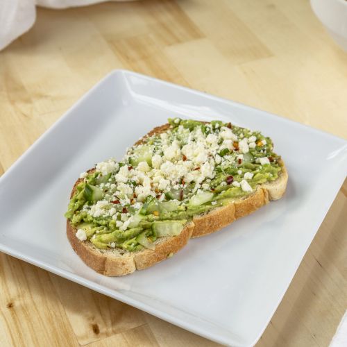 A piece of avocado toast topped with feta cheese on a white square plate, next to a fork and a bowl of soup on a wooden table.