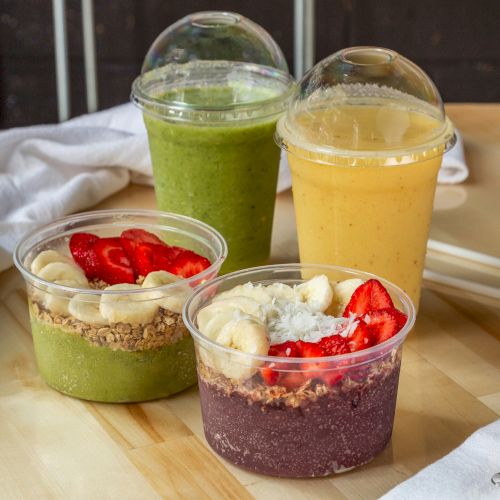 The image shows two smoothie bowls topped with fruits and granola, alongside two smoothies, one green and one yellow, on a table with a cloth and spoon.