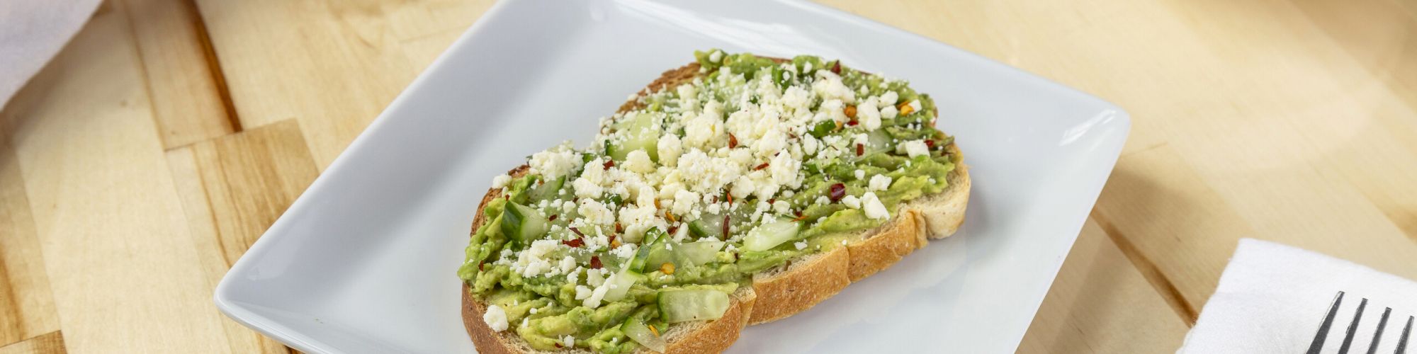 A slice of bread topped with mashed avocado and crumbled cheese is on a white square plate, with a fork and bowl in the background.