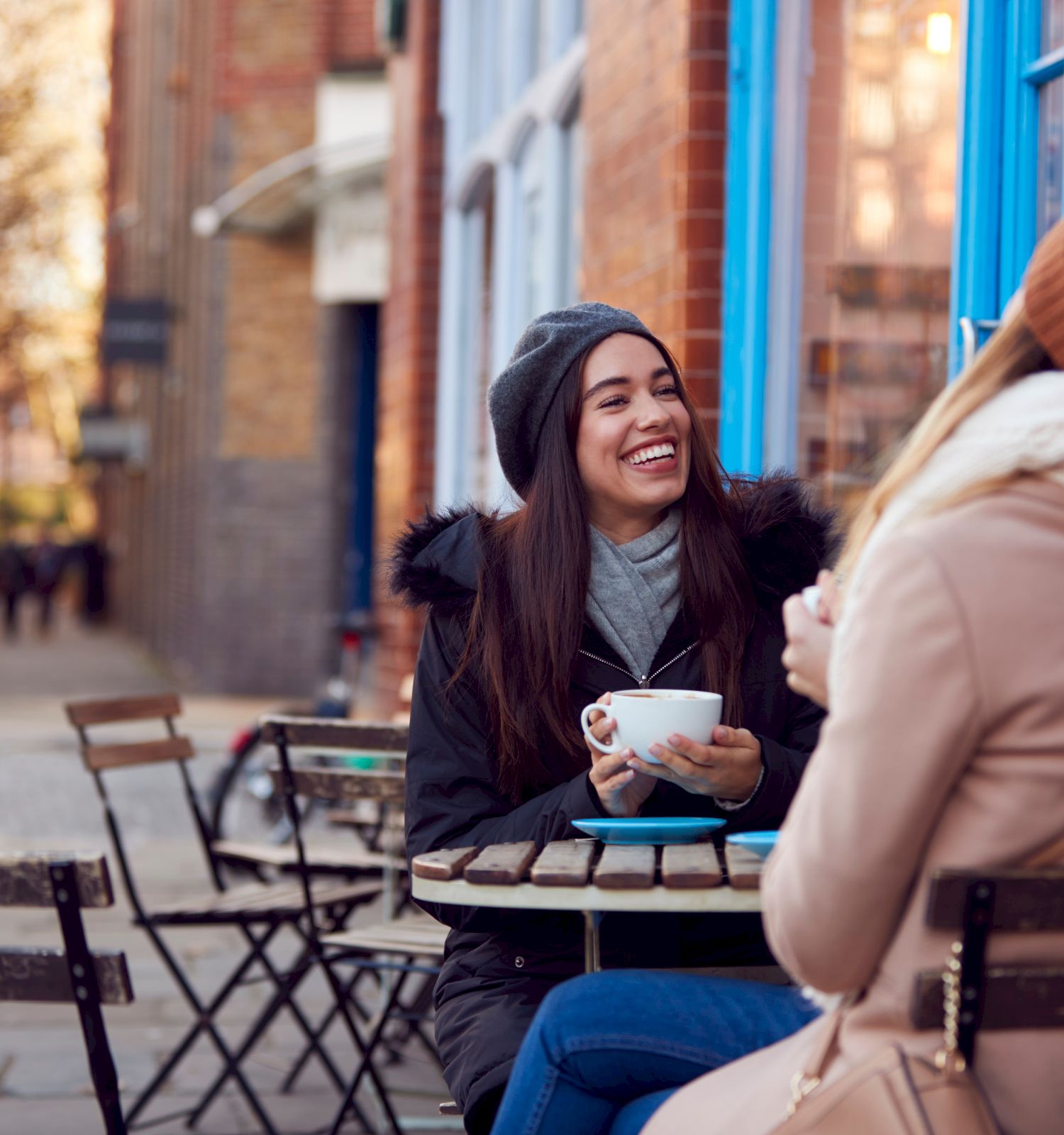Two women are sitting outside at a cafe table, enjoying beverages and smiling at each other. They are dressed warmly in winter clothing.