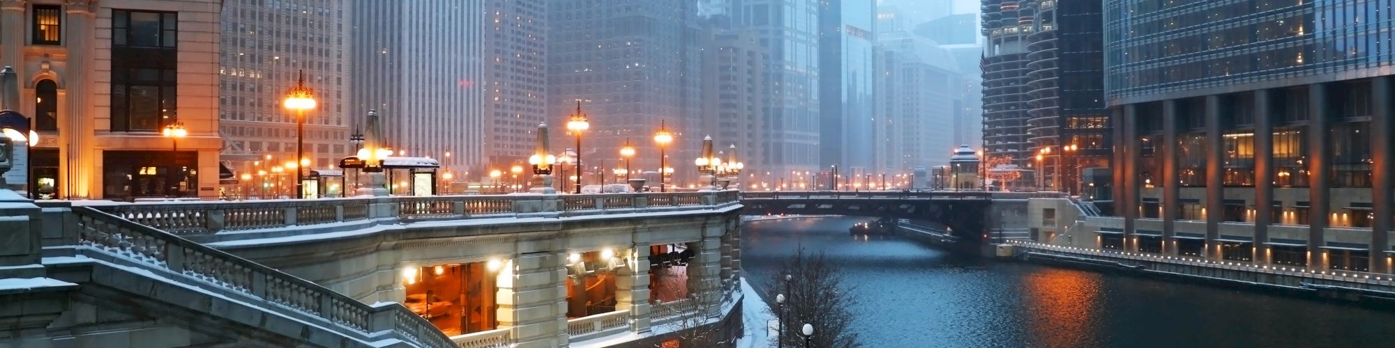 A snowy cityscape with tall buildings and a river flanked by a walkway. Streetlights glow in the early evening, casting reflections on the water.