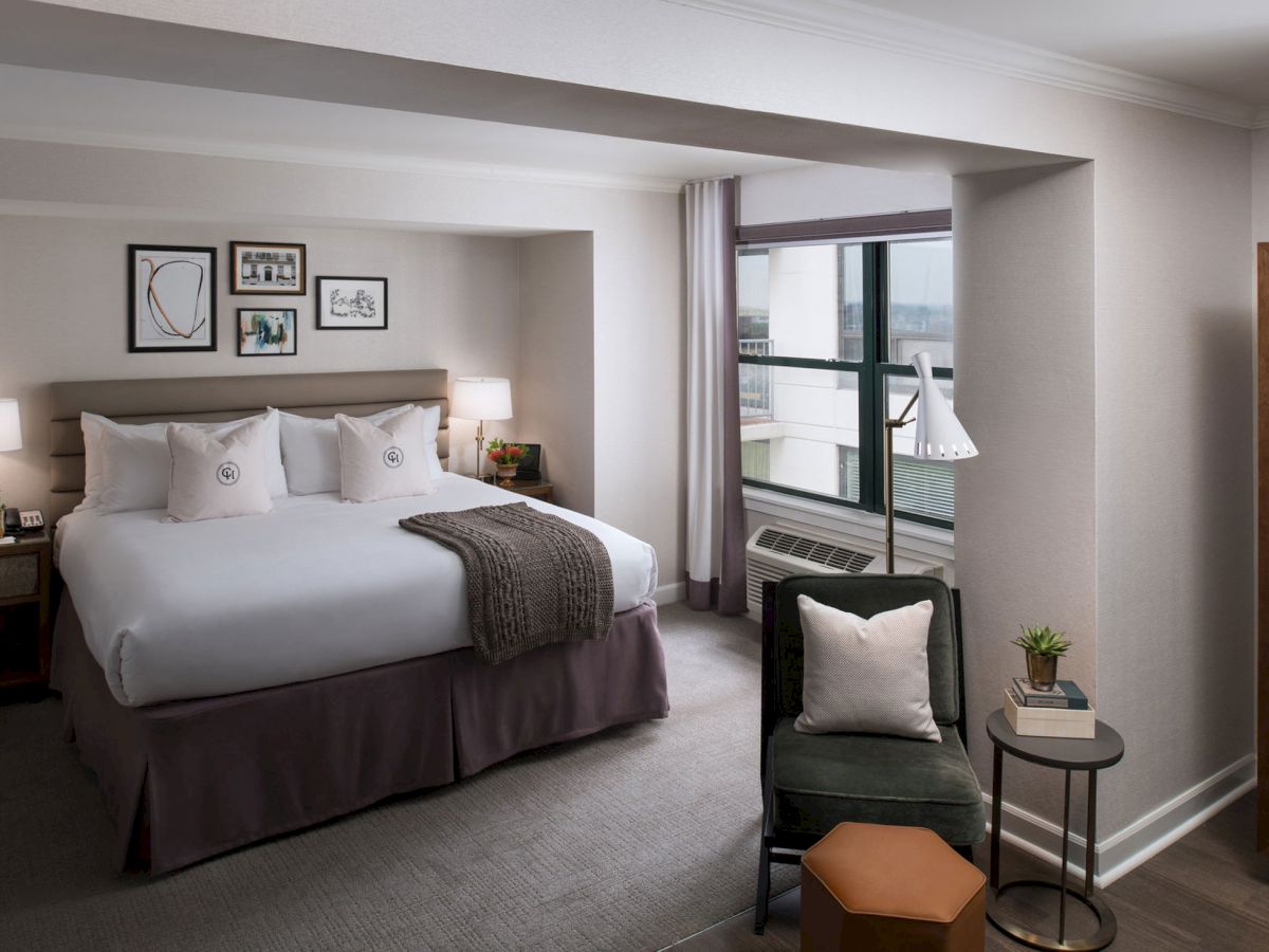 A hotel room with a large bed, neutral tones, nightstands with lamps, a chair and small table by the window, and framed art on the wall.