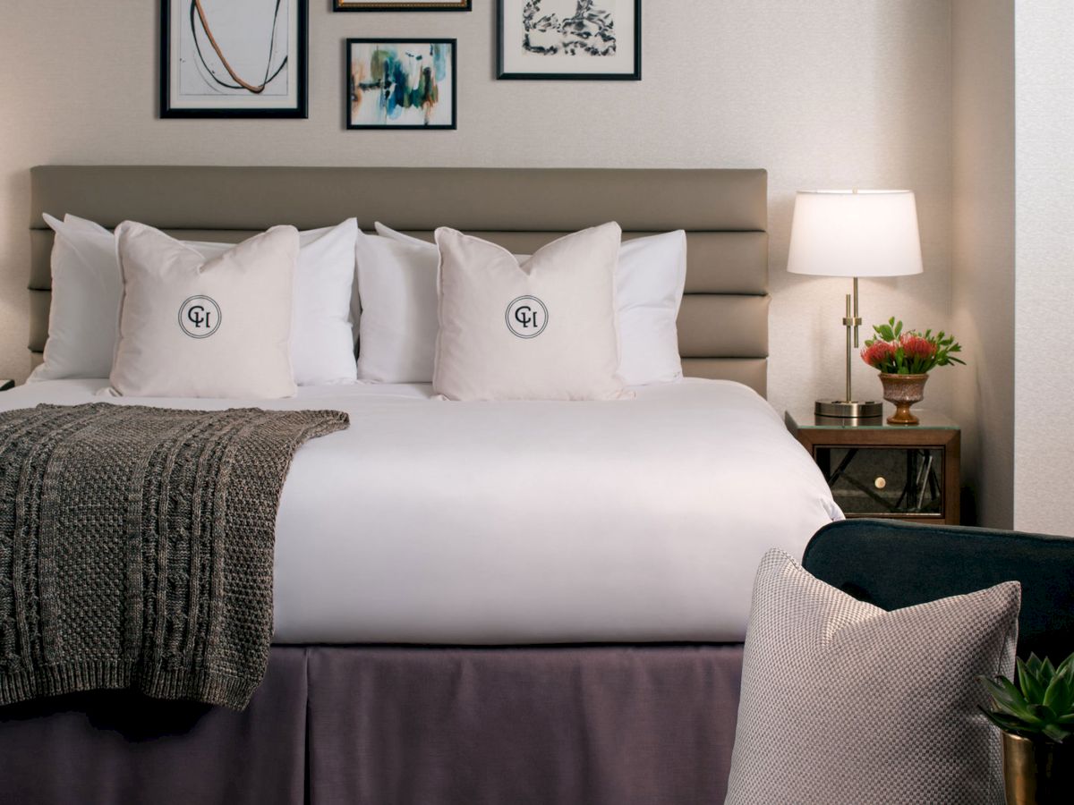 A stylish hotel bedroom with a large bed, white linens, framed art above, a nightstand lamp, and a cozy seating area by the foot of the bed.