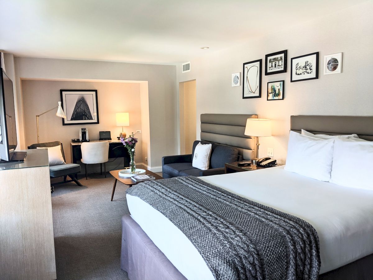 A modern hotel room with a large bed, gray headboard, throw blanket, and white bedding, plus a seating area, desk, and wall art.