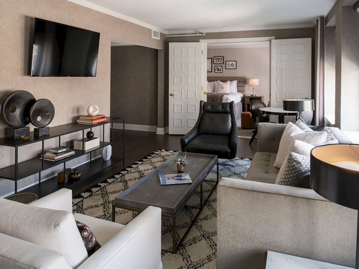 A stylish living room with a wall-mounted TV, open shelving, a patterned rug, white sofas, a coffee table, and a view into a bright bedroom.