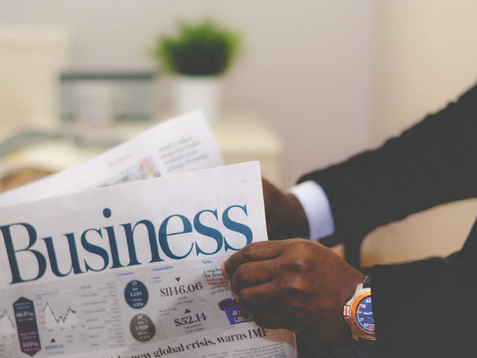 A person in a suit reads a printed business newspaper on a desk, focusing on market charts and headlines, in a professional office setting.