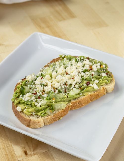 Sliced baguette topped with mashed avocado, chopped greens, feta crumbles, and herbs on a white plate, with a bowl of soup in the background.