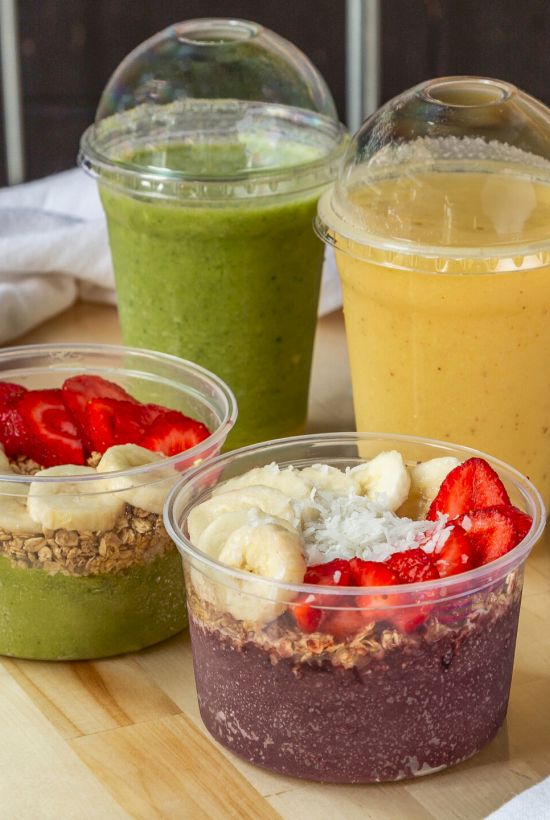 A trio of colorful smoothie bowls (green, yellow, and purple) topped with strawberries and cream, with two smoothies in cups behind them.
