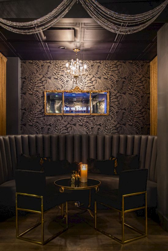 A chic, dimly lit lounge with a dark patterned wall, a curved sofa, a round table set for four, decorative drapes, and tropical plants in pots, candlelight.