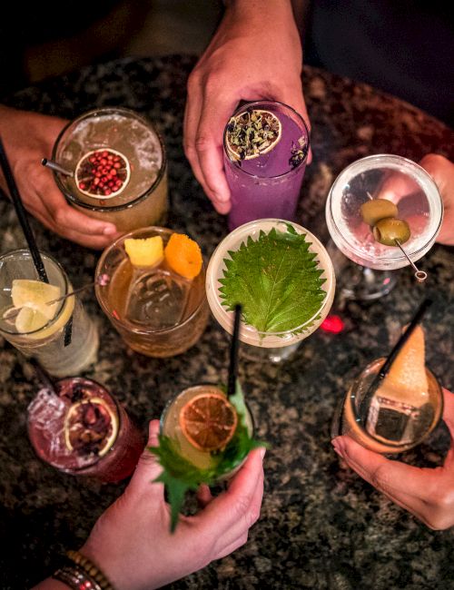 Six colorful cocktails in round glasses, garnished with fruit and herbs, around a central lime and ice.