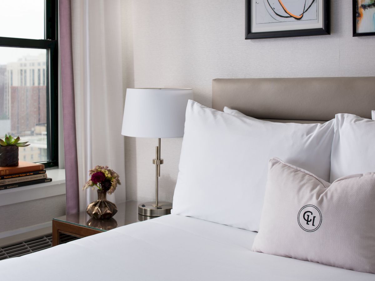 A tidy hotel-style bedroom: neatly made bed with white linens and pillows, a bedside lamp, small stack of books, plant on the windowsill, and framed art above the headboard.
