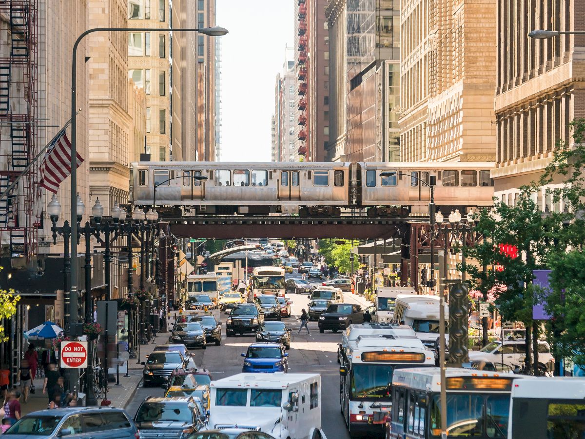 A busy city street with cars, buses, tall buildings, and an elevated train passing overhead, suggesting a bustling urban transit scene.