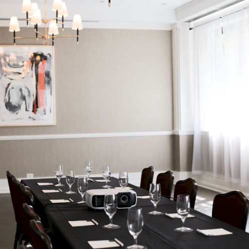 A long dining table set with wine glasses, napkins, and cutlery in a softly lit, modern room with wall art and white curtains.