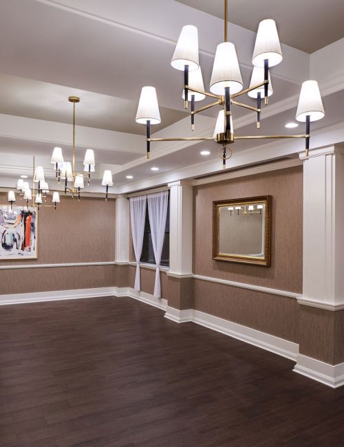 Elegant banquet or event room with warm brown walls, white trim, chandeliers, and sheer draped windows&mdash;ready for gatherings.