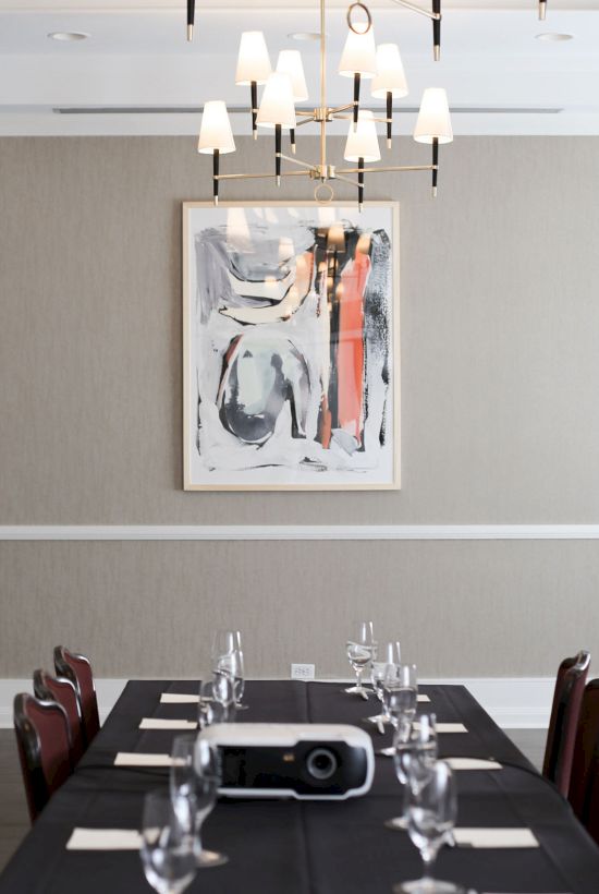 A neatly set conference room with a long table, chairs, glassware, a projector, and a framed abstract painting on a beige wall.