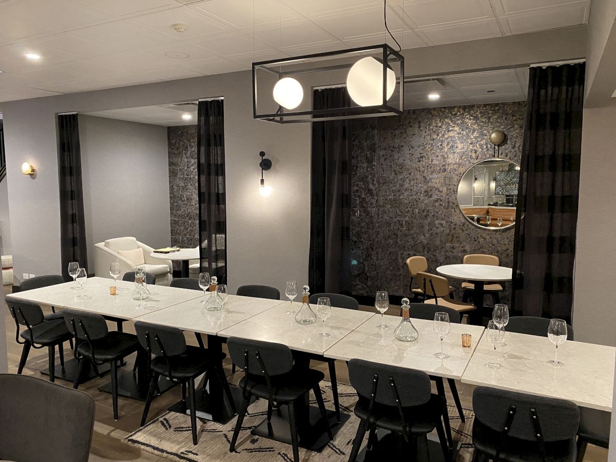 A long white dining table with black chairs in a modern restaurant, glassware set, stylish lighting, and a textured wall with a round mirror.