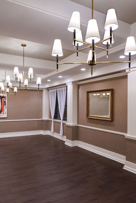 A spacious, well-lit banquet-style room with chandeliers, brown wainscoting, white trim, dark wood floors, and framed art by the windows.