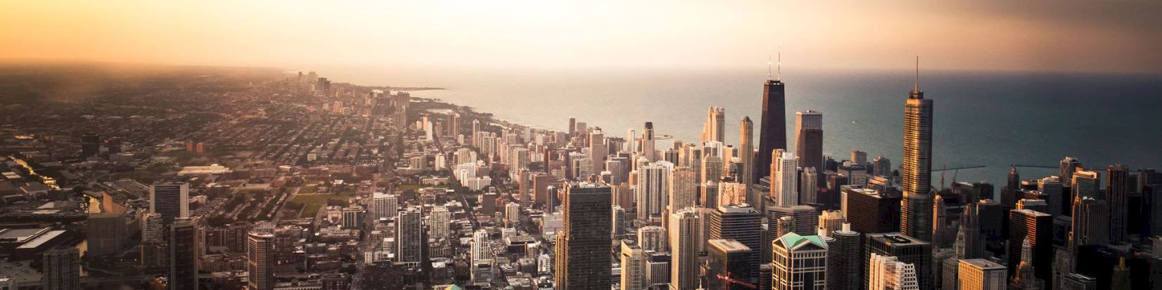 A panoramic city skyline at dusk with tall buildings near the water, glowing with warm sunlight and a calm horizon beyond.