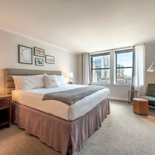 A bright hotel or apartment bedroom with a large bed, nightstands, wood furniture, a sofa, and a sunny windowed view with neutral decor.