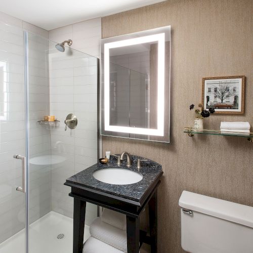 A compact bathroom with a dark vanity, white sink, backlit mirror, glass shower enclosure, beige tiled walls, and a small chair by a towel rack.