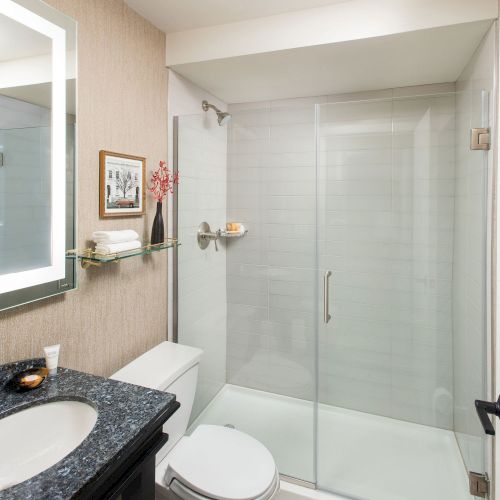 A modern bathroom with a glass shower enclosure, white tile, a dark vanity with a round mirror, and a toilet between.