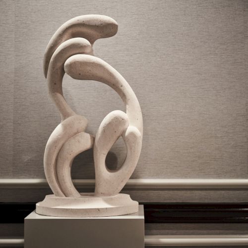 A decorative white ceramic sculpture shaped like a stylized, abstract figure sits on a wooden shelf against a dark, textured wall.