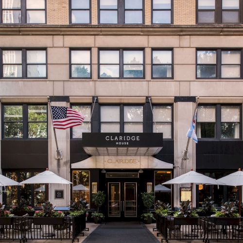 A grand hotel facade with a wrought-iron awning, cream stone, and leafy outdoor seating by black umbrellas. Top it at 140 characters, always ending the sentence.