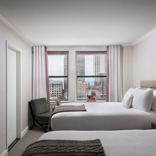 A bright, modern hotel bedroom with a large bed, neutral tones, two nightstands, a seating area, a window view, and warm ambient lighting.