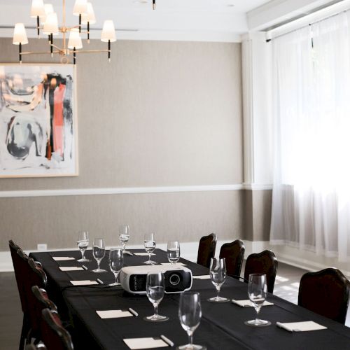 A chic dining room set for a formal meal: a long black table, wine glasses, napkins, silverware, chairs, soft lighting, and modern artwork on the wall.