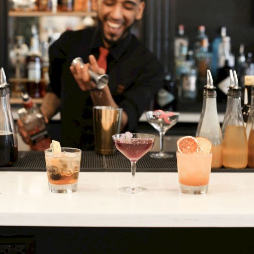 Bartender smiling and shaking a cocktail shaker behind a bar with colorful drinks on the counter, ready to pour.