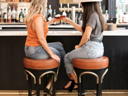 Two women sit on barstools, clinking glasses, at a bar with various bottles in the background. One wears a hat, both in casual attire.